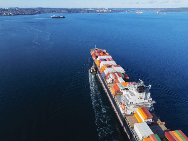Aerial view of a  ship nearing port with the assistance of a tugboat, illustrative of wine trade tariffs.